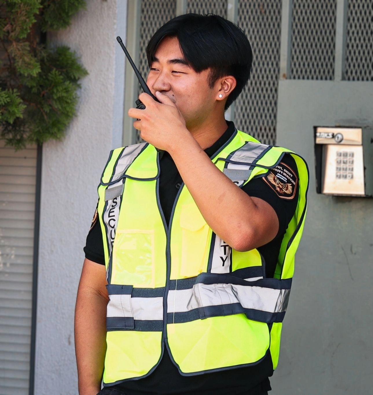 security guard standing by the entrance of a building wearing a reflective vest talking into a radio